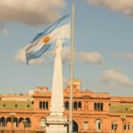 Bandera de Argentina flameando al viento junto a la Pirámide de Mayo, con la Casa Rosada de fondo en un día soleado.