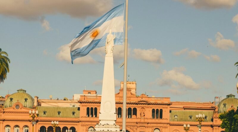Bandera de Argentina flameando al viento junto a la Pirámide de Mayo, con la Casa Rosada de fondo en un día soleado.