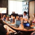 Hombre presenta ante grupo de personas sentadas en mesa de conferencia con laptops y bolsas de regalo.