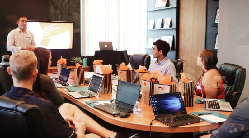 Hombre presenta ante grupo de personas sentadas en mesa de conferencia con laptops y bolsas de regalo.