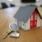 Maqueta de casa en miniatura de color blanco y rojo con techo gris, acompañada de un juego de llaves con llavero en forma de casa sobre una mesa de madera.