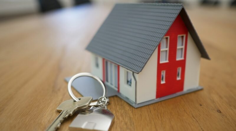 Maqueta de casa en miniatura de color blanco y rojo con techo gris, acompañada de un juego de llaves con llavero en forma de casa sobre una mesa de madera.