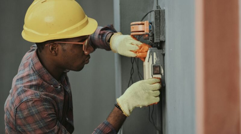 Electricista con casco amarillo, gafas y guantes de trabajo instalando o revisando un cuadro eléctrico en una pared gris.