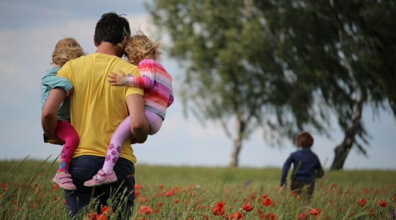 hombre camina en un parque abierto en el que carga a dos hijas y más adelante corre un hijo varón.