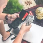 Un hombre saca billetes de dólares de su billetera negra sobre una mesa de madera con una laptop y un globo terráqueo.