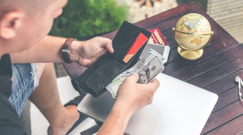 Un hombre saca billetes de dólares de su billetera negra sobre una mesa de madera con una laptop y un globo terráqueo.